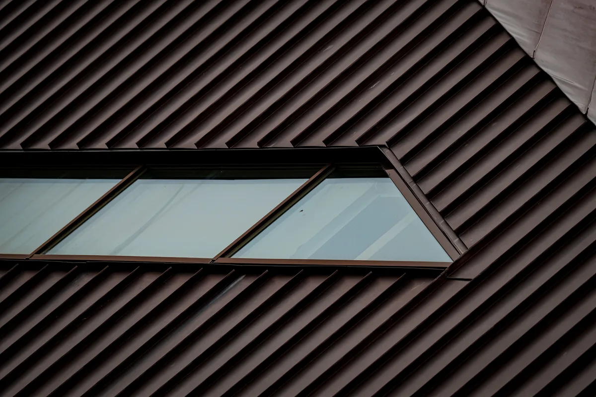 A dark raised-seam metal roof seen up close, parallel metal seams with a narrow skylight set between two panels
